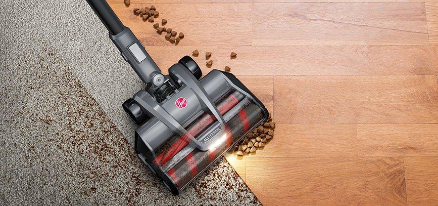 Hoover vacuum cleaner demonstrating its ability to clean both carpet and hard floor types with two brush rolls. The vacuum is shown picking up pet food from both surfaces, highlighting its versatility