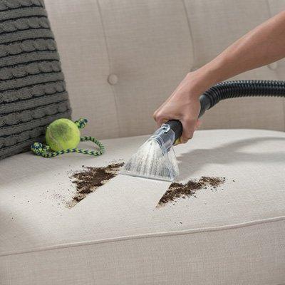 Someone using a Hoover upholstery brush to clean dirt from a couch demonstrating removal of stains