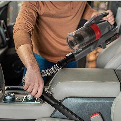 A person using a Hoover emerge pet vacuum cleaner to clean the front seat of the car, showcasing the vacuum's versatility and ability to reach tight spaces with its flexible hose and specialized attachments.