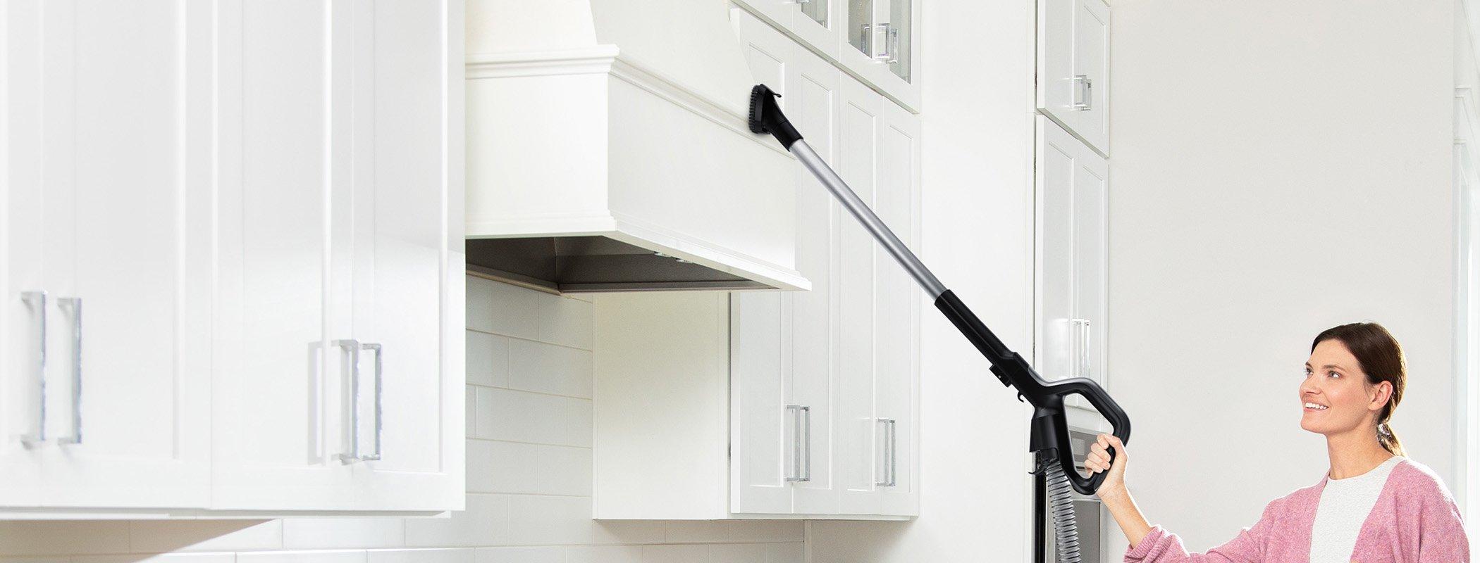 Woman using Hoover vacuum cleaner with extended reach wand to clean high kitchen cabinets, highlighting versatility and ease of use