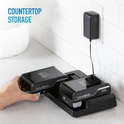 Hand placing a Hoover ONEPWR lithium ion battery into a countertop charging station with another battery already stored and charging, featuring a wall-mounted power adapter.