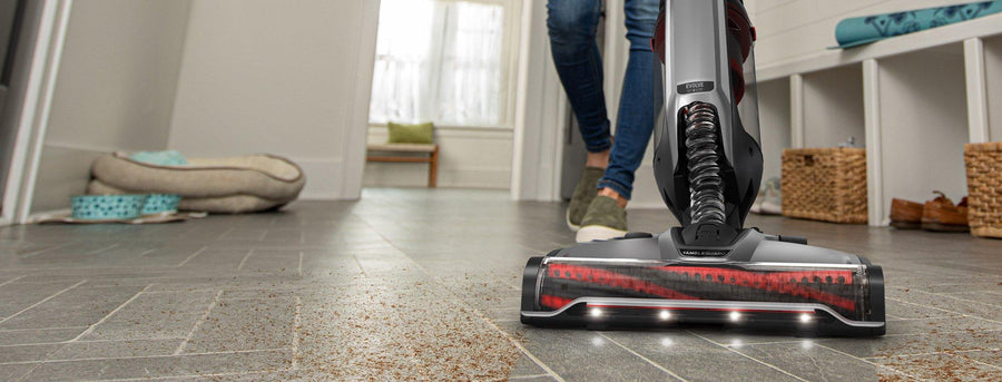 Close-up view of a Hoover vacuum cleaner with LED lights, cleaning a carpet, highlighting its effective dirt pickup as a person walks in the background. 