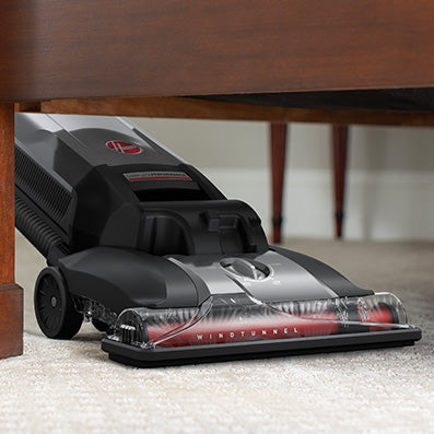 Close up of a Hoover vacuum cleaner cleaning under furniture, showcasing it's flexibility and windtunnel technology for effective dust and debris removal.