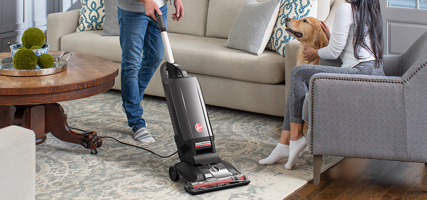 Person vacuuming a living room carpet with a Hoover vacuum cleaner, while a woman and dog sit on a couch watching.