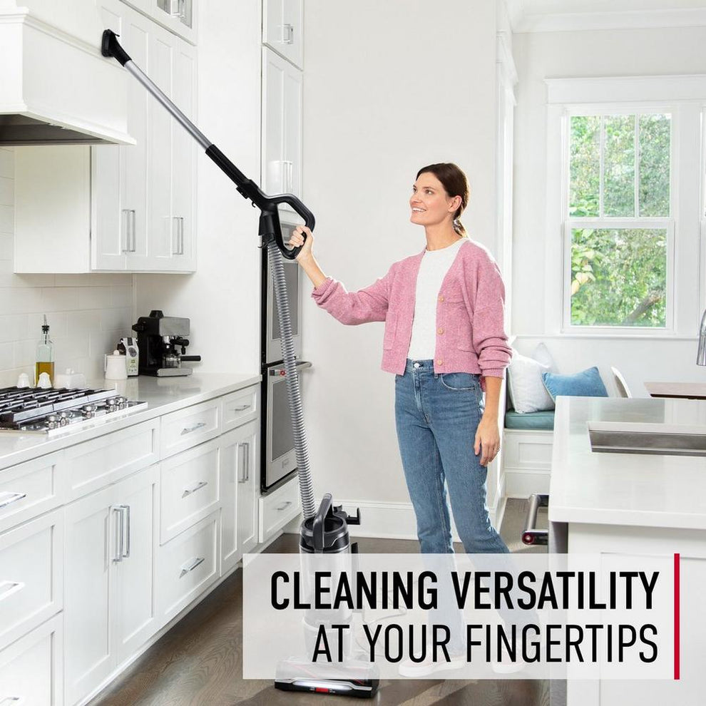 Woman using Hoover vacuum cleaner with an extension wand to clean high kitchen cabinets, with text "cleaning versatility at your fingertips," highlighting ease of use and flexibility.