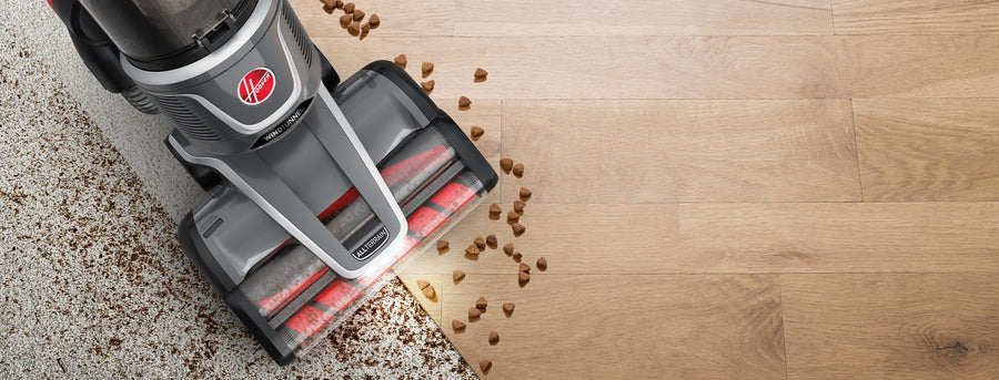 Top down view of a Hoover windtunnel vacuum cleaner seamlessly transitioning from carpet to hardwood floor, effectively picking up scattered pet food and dirt