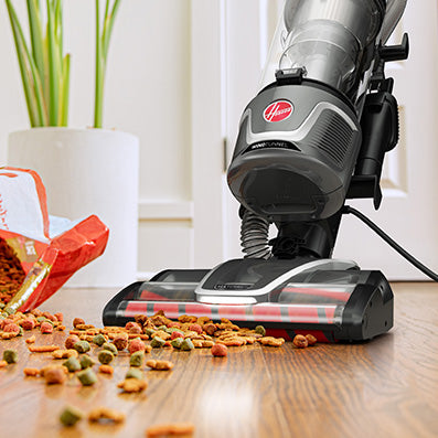 Hoover upright vacuum cleaner, ideal for pet owners, is shown in action cleaning spilled dog food on a hardwood floor.