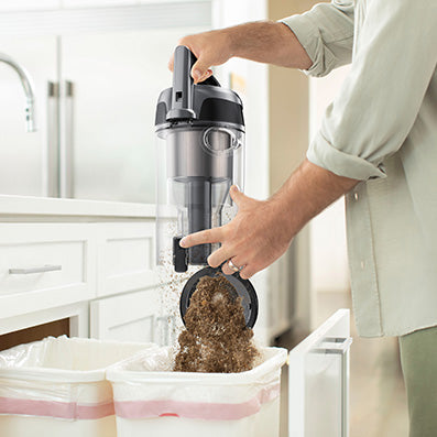 Close up of a person emptying the dirt cup of a Hoover windtunnel vacuum cleaner into a trash can, emphasizing the vacuum's easy maintenance