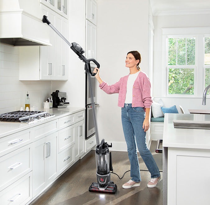 Woman using a Hoover upright vacuum cleaner with the extended hose attachment and 2 in 1 upholstery tool to clean her hard to reach kitchen cabinets and kitchen hood